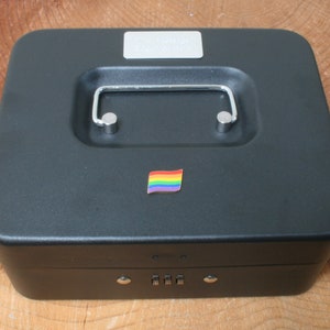 May include: Black metal cash box with a combination lock and a rainbow flag sticker. The cash box has a handle on the top.