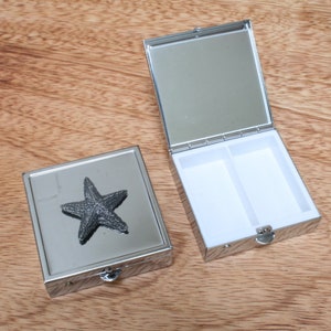 May include: A silver pill box with a starfish design on the lid. The box has two compartments inside.
