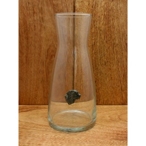 Può includere: Una caraffa di vetro trasparente con un tappo di metallo a forma di testa di cane. La caraffa è su una superficie di legno.