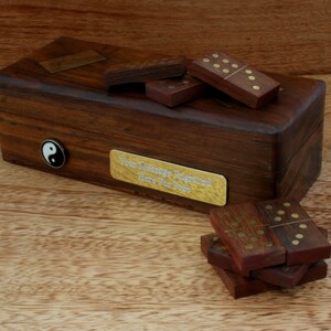 Puede incluir: Una caja de madera con una incrustación de latón y una placa de latón que dice "Su mensaje grabado aquí gratis". La caja contiene un juego de dominós de madera con puntos de latón.