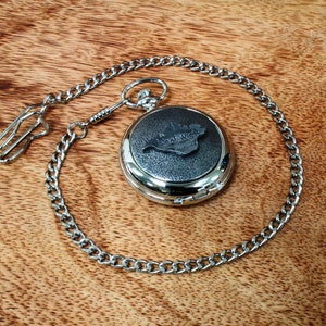 May include: A silver pocket watch with a black face and a chain. The watch face has an inscription that reads "Isle of Wight".