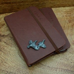 May include: Two brown leather bound notebooks with the word "NOTES" embossed on the cover. The notebooks are stacked on top of each other and have a silver metal bird charm attached to the top notebook.