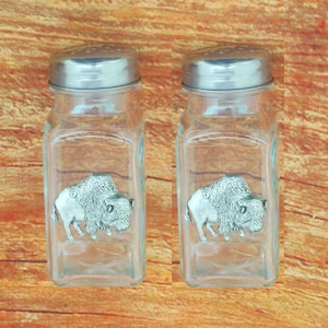 May include: A set of two glass salt and pepper shakers with pewter buffalo figurines on the front. The shakers are clear glass with silver lids.