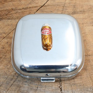 Puede incluir: Una caja de cigarros de metal plateado con una tapa con bisagras. La tapa presenta un cigarro decorativo de color dorado con una banda roja que dice "Cuban".
