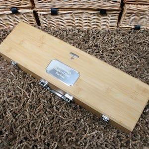 May include: A light-colored bamboo wine box with silver hardware and a silver plaque that reads "Your Message Engraved Here For Free." The box is rectangular and has a handle. The background includes wicker baskets.
