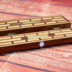 May include: A wooden cribbage board with a brass inlay and a small blue and gold emblem. The board is closed and sitting on a wooden surface.