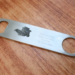 May include: A silver bottle opener with a small metal combine harvester design. The opener has a space for engraving with the text "Your Message Engraved Here For Free Up To 4 Lines".