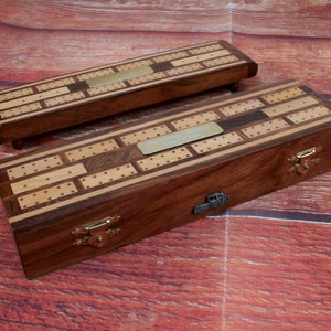May include: Two wooden cribbage boards with brass hardware and a brass plaque for engraving. The boards are made of light and dark wood with a patterned inlay.