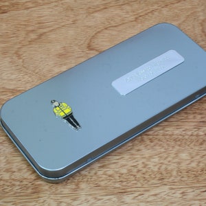 May include: A silver metal tin with a yellow and black cartoon image of a person wearing a yellow safety vest. The tin has a silver label with the text "To a Amazing Teacher" printed on it.
