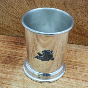 May include: A silver-colored metal cup with a detailed emblem of a combine harvester. The cup has a polished interior and a textured exterior, with a wide base and a rolled rim. The cup is likely for decorative or collectible purposes.