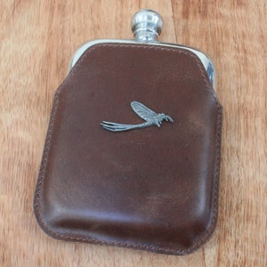 May include: A brown leather flask with a silver metal top and a silver metal dragonfly design on the front.
