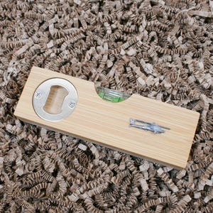 Könnte beinhalten: Ein Flaschenöffner und Wasserwaage aus Bambus. Das Werkzeug hat einen silbernen Flaschenöffner, eine Libelle und ein kleines silbernes Flugzeugemblem. Das Werkzeug ist rechteckig und aus hellem Holz gefertigt.