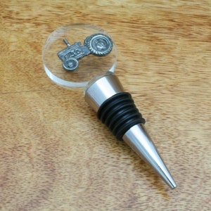 May include: A silver metal bottle stopper with a black rubber seal. The top of the stopper features a clear acrylic circle with a silver metal tractor design.