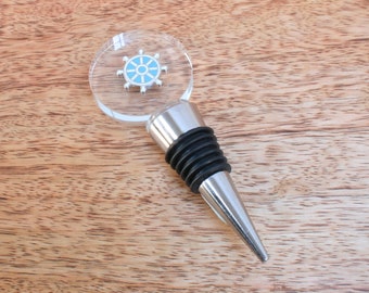 May include: A wine bottle stopper with a clear, circular top featuring a blue and silver ship's wheel design. The stopper has a silver metal base and a black rubber seal. The product is set against a wooden background.