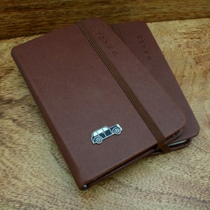 May include: Two brown leather-bound notebooks with the word "NOTES" embossed on the covers. The top notebook has a small silver car charm attached.