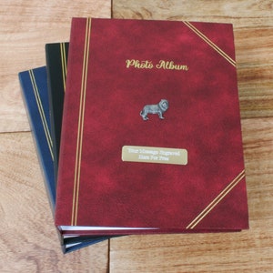 Puede incluir: Tres álbumes de fotos con cubiertas roja, azul y negra. El álbum rojo tiene un emblema de león dorado y el texto "Photo Album" en la parte delantera. El texto "Su mensaje grabado aquí gratis" está en una placa plateada en la parte delantera del álbum rojo.