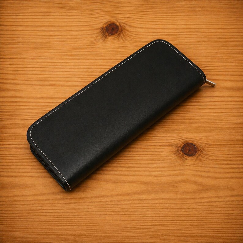 K&ouml;nnte beinhalten: Ein schwarzes Leder-Etui mit Rei&szlig;verschluss und wei&szlig;en N&auml;hten an den Kanten. Das Etui ist rechteckig und scheint geschlossen zu sein. Der Rei&szlig;verschluss ist silberfarben. Das Etui liegt auf einer Holzoberfl&auml;che.