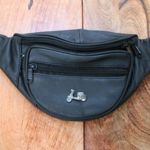 Puede incluir: Riñonera de cuero negro con un dije de scooter plateado. La riñonera tiene dos compartimentos con cremallera y un lazo para el cinturón.