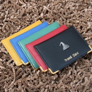 May include: A stack of colorful travel card holders in yellow, blue, green, red, and black. The black holder has a silver hat emblem and the words "Travel Card" in gold. Each holder has gold corner accents.