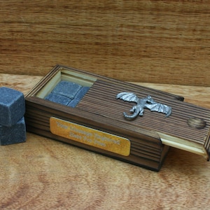 May include: Wooden box with a sliding lid, featuring a silver dragon design. The box contains a set of gray whiskey stones and a gold plaque with the text "Your Mileage May Vary Here's to the Journey".