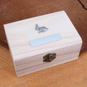 Könnte beinhalten: Eine kleine Holzkiste mit einem Scharnierdeckel und einem Metallverschluss. Die Kiste hat eine silberne Hasenfigur auf der Oberseite und eine silberne Plakette mit dem Text "The Adventures of a Little Bunny" auf der Vorderseite.