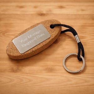 Könnte beinhalten: Ein ovaler Schlüsselanhänger aus Kork mit einer silbernen Metallplatte, auf der "Your Message Engraved Here" steht. Er hat eine schwarze Kordel und einen silbernen Schlüsselring. Der Kork hat ein natürliches, strukturiertes Aussehen.