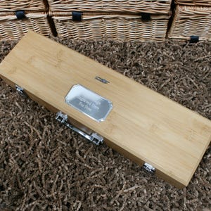 May include: A rectangular bamboo box with a silver metal handle and clasps. A silver plaque on the box reads "Engraved Here For Free". The box is on a brown textured surface, with several wicker baskets in the background.