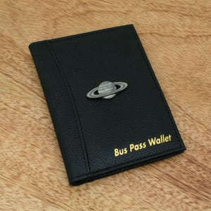 Peut inclure: Porte-carte de bus noir avec un emblème de planète argenté. Le portefeuille a une surface texturée et des lettres dorées qui disent "Bus Pass Wallet". Le portefeuille est fermé et repose sur une surface en bois.