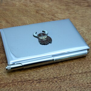 Op de afbeelding: Een zilveren metalen sigarettenkoker met een Vikinghoofd ontwerp. De koker heeft een scharnierend deksel en een klein, zilveren metalen slot.