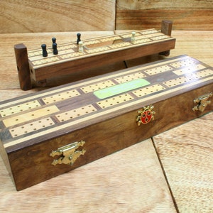 Op de afbeelding: Een houten cribbage-bord met een groen label waarop "Cribbage Board" staat. Het bord heeft een bruine en lichte houtkleur en heeft messing scharnieren en een lieveheersbeestje-ontwerp aan de zijkant. Het bord is gesloten en ligt op een houten oppervlak.