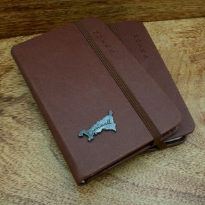 May include: Two brown leather notebooks with elastic closures. The top notebook has a metal Cornwall map charm attached to the front cover. Both notebooks have the word "NOTES" embossed on the front cover.