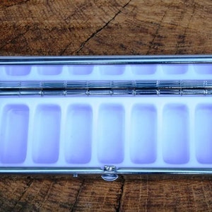 May include: A silver pill box with a hinged lid and seven compartments. The compartments are lined with a light purple material.