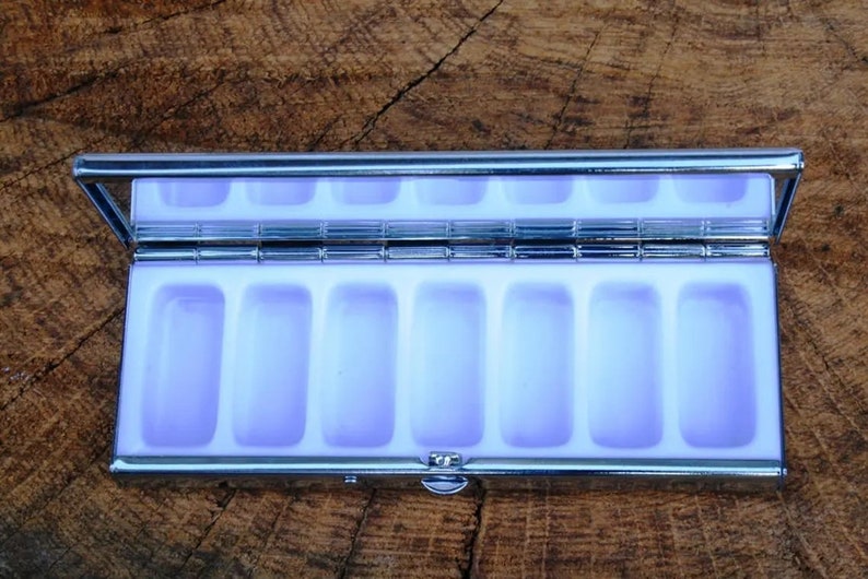May include: A silver pill box with a hinged lid and seven compartments. The compartments are lined with a light purple material.