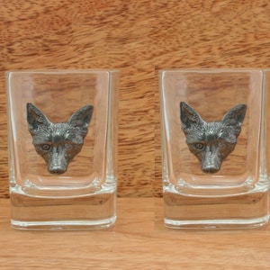 Puede incluir: Dos vasos de chupito de vidrio transparente con un diseño de cabeza de zorro de peltre. La cabeza de zorro es de color gris oscuro.