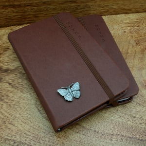 May include: Two brown leather-bound notebooks with elastic closures. The top notebook has a silver butterfly charm attached. Both notebooks have the word "NOTES" embossed on the front cover.