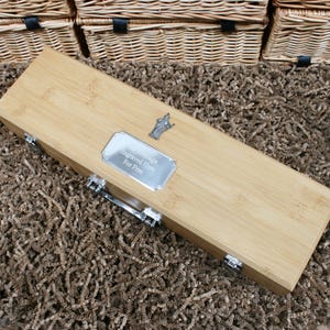 Puede incluir: Una caja de vino rectangular de bambú con herrajes plateados y una placa plateada que dice "Your Message Engraved Here For Free". La caja está cerrada y descansa sobre una superficie marrón texturizada.