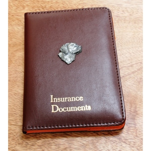 May include: A brown leather insurance document holder with a silver dog head ornament and the words "Insurance Documents" in gold lettering.