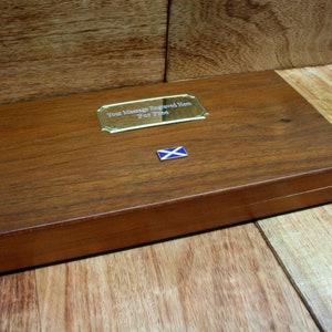 Puede incluir: Una caja rectangular de madera con un acabado pulido. Una placa dorada con el texto "Your Message Engraved Here For Free" está fijada en la parte superior. También hay un pequeño emblema de la bandera escocesa. La caja está sobre una superficie de madera.