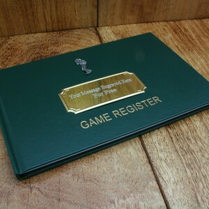 Könnte beinhalten: Ein grünes, ledergebundenes Buch mit einer goldenen Plakette, auf der "Ihre Botschaft hier kostenlos eingraviert" steht, und den Worten "SPIELREGISTRIERUNG" in goldenen Lettern.