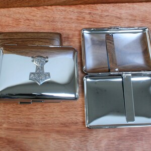 May include: A silver cigarette case with a raised design of a hammer and a face on the front. The case is open and the inside is also silver.