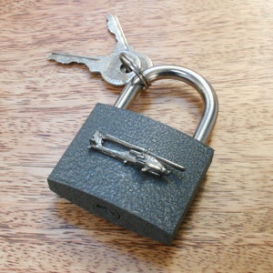 Könnte beinhalten: Ein graues Vorhängeschloss mit einem silbernen Hubschrauber-Anhänger. Das Vorhängeschloss liegt auf einer Holzoberfläche.