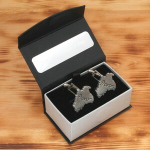 Peut inclure: Une boîte cadeau noire avec un intérieur blanc, contenant deux boutons de manchette en argent en forme de contour du Devon, en Angleterre. Chaque bouton de manchette porte le mot "Devon" gravé dessus.