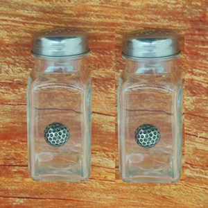 Op de afbeelding: Twee glazen zout- en peperstrooiers met zilveren deksels en een golfbalontwerp aan de voorkant.