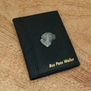 May include: Black bus pass wallet with a silver dog head emblem. The wallet has the words "Bus Pass Wallet" in gold lettering. The wallet is made of a textured material and is on a wooden surface.