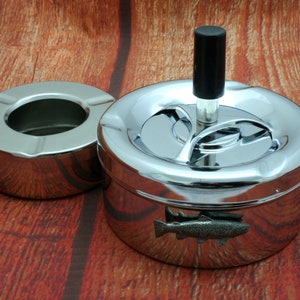 May include: A chrome ashtray with a rotating lid and a fish design. The ashtray has a black knob on the top and a separate, smaller ashtray on the side.