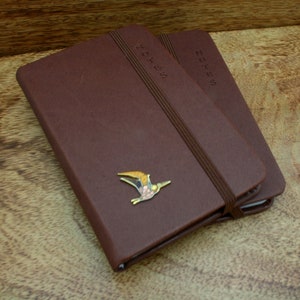 May include: Two brown leather bound notebooks with the word "NOTES" embossed on the front. The top notebook has a small gold bird pin attached to the front cover.