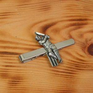 May include: A silver tie clip featuring a cricket player in action. The player is holding a bat and is in a batting stance. The tie clip is attached to a silver bar.
