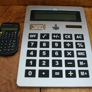 May include: A silver calculator with a black display and black buttons. The calculator has a square, silver Masonic symbol on the top. The text "Your Message Engraved Here For FREE" is on a silver tag on the calculator.