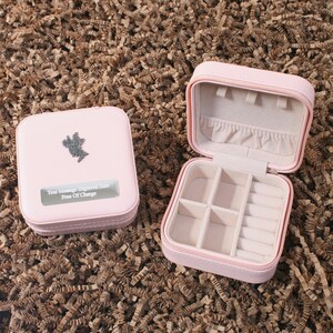 May include: Two pink jewelry boxes, one open and one closed, on a brown surface. The closed box has a silver emblem and the text "Your Message Engraved Here Free Of Charge". The open box reveals compartments.