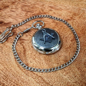 Op de afbeelding: Zilveren zakhorloge met een ketting. Het horloge heeft een zilveren vrijmetselaars symbool met een passer en een winkelhaak.